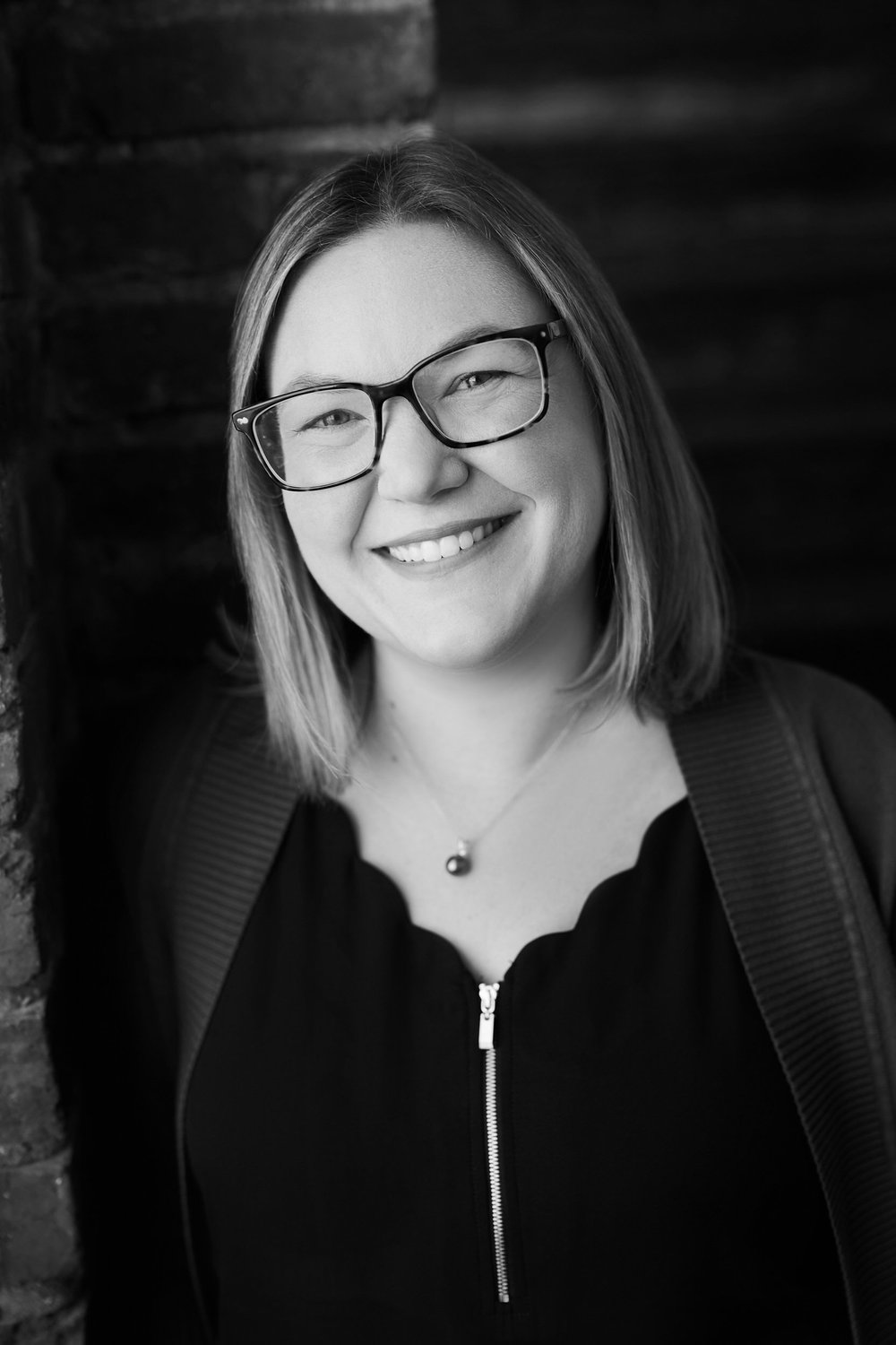 Elena Hutchison, the founder of Up/Right Analyst Relations and former Chief Strategy Officer at Medallia, stands smiling in glasses and shoulder-length hair, wearing a zip-up jacket and necklace, against a brick wall.